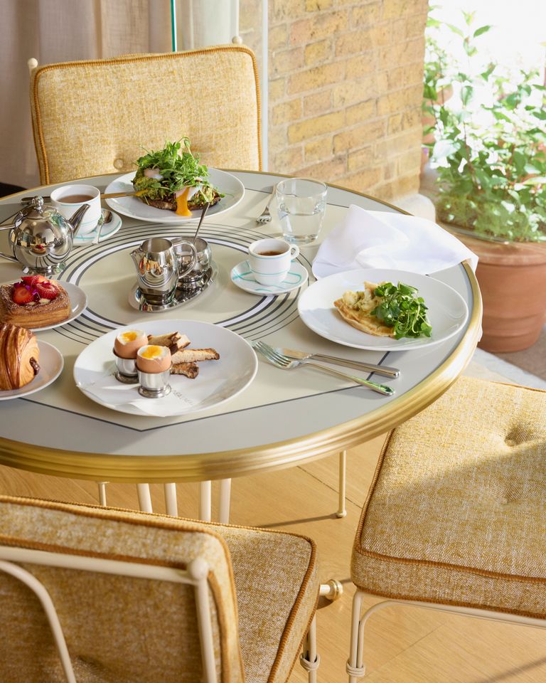 طاولة دائرية مغطاة بكloth أبيض تحتوي على أطباق فطور لذيذة، بما في ذلك بيض مقلي، ساندوتش خبز محمص، وكعكة محشوة بالتوت، مع قهوة وشاي في أكواب صغيرة