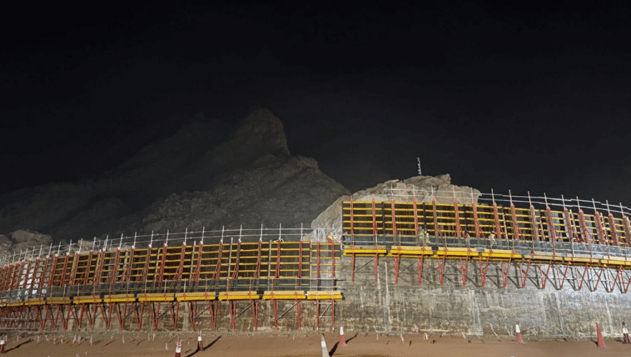 A nighttime construction site featuring colorful scaffolding supporting structural elements, with a large mountain in the background.