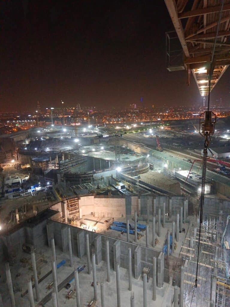 An image of an ongoing construction project at night, showcasing a massive architectural structure under development, illuminated by bright worklights. The building features tiered layers and distinctive architectural elements, with cranes actively lifting materials and assembling components.
