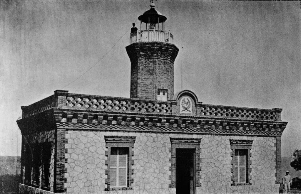 An old lighthouse built with natural stone, featuring a cylindrical tower and an ornate balcony, with a person standing at the top.