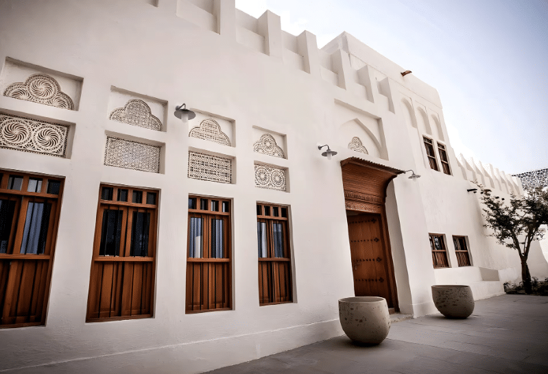 مبنى أبيض تقليدي يُظهر نوافذ خشبية مزخرفة وประตًا كبيرًا مصنوعًا من الخشب، مع زخارف هندسية على الجدران تضيف لمسة فنية، ونباتات صغيرة في حاويات خرسانية أمام المدخل.