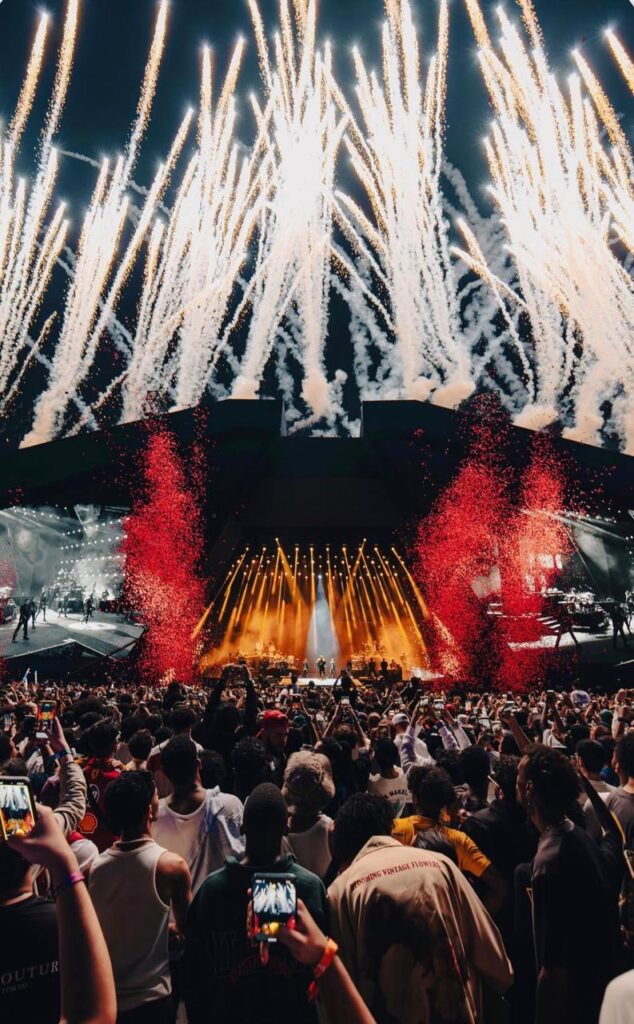 A large-scale music concert with fireworks lighting up the sky, as the crowd engages with the performance.