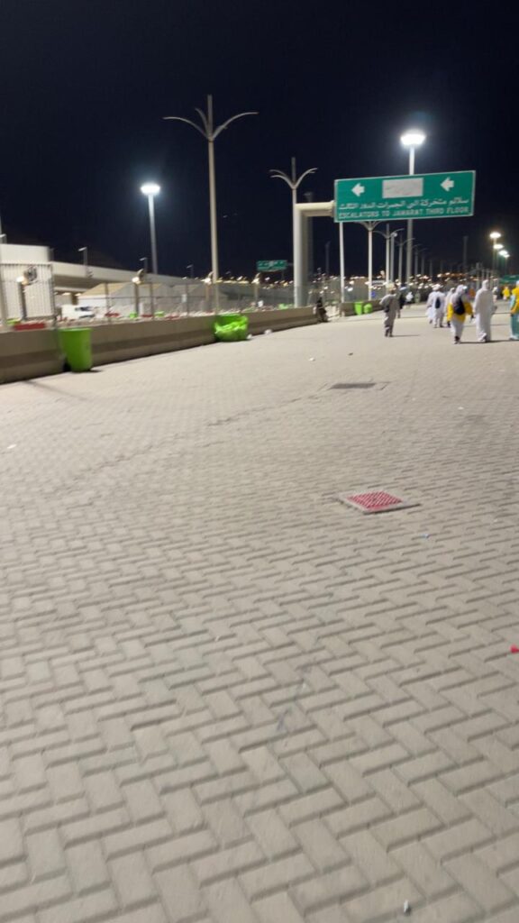 A night street in the Hajj area featuring a directional sign and a pile of waste near green bins.