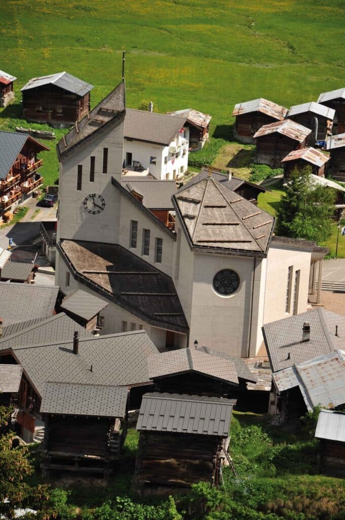 A white church with a minimalist architectural design, situated in a small Swiss village surrounded by traditional wooden houses and expansive green fields.