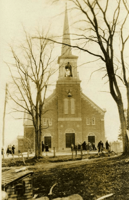 An old black-and-white photograph of a small, historic church with a tall, slender steeple and a cross at the top. The building is constructed from stone or brick, featuring simple architectural details such as arched windows and a central entrance. A few people are gathered outside, standing near the church and along a partially unpaved road. Trees surround the area, adding to the serene and rustic atmosphere.