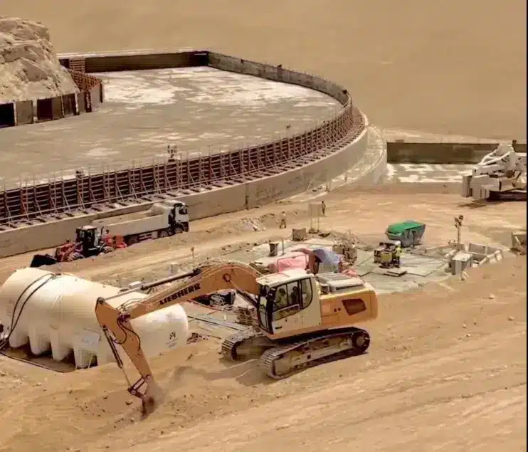 A construction site in the desert featuring a large circular building under construction, with heavy machinery operating on-site.