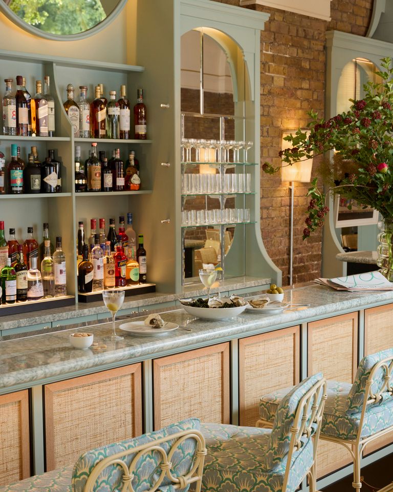 A bar with shelves stocked with various alcoholic beverages, small plates of food on the counter, and warm lighting that highlights the brick walls and decorative furniture