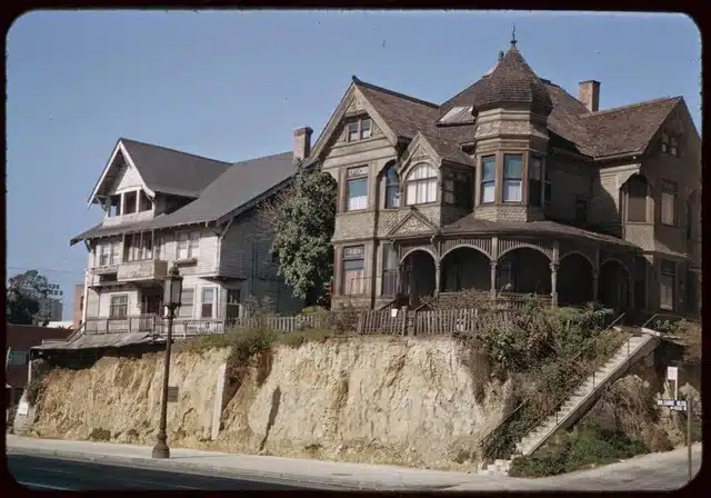 A grand Victorian house with elaborate architectural features, including balconies and decorative windows. The house is located on a hillside, surrounded by trees and plants, with stairs leading to the main entrance.