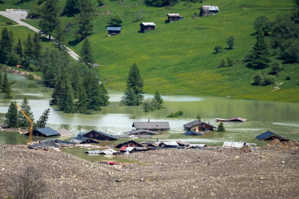 A view of a small Swiss village showing the aftermath of a natural disaster, where water has submerged most of the houses and fields due to a glacial collapse and debris accumulation.