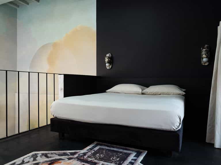 A modern and artistic bedroom featuring a white bed centered against a dark black wall with a painted mural of a sunset on the adjacent white wall. Two vibrant wall-mounted lamps add an artistic touch, complemented by a geometric-patterned rug. A wooden-framed window adds a traditional element, while natural light enhances the room's beauty.
