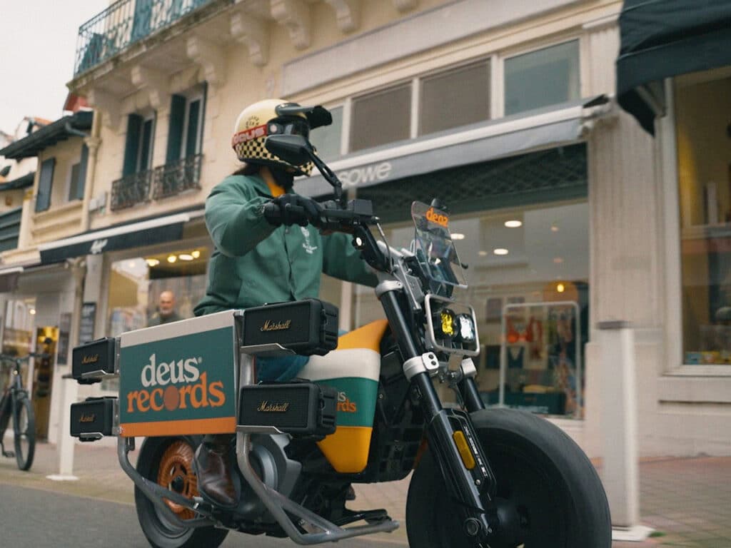 Person riding the BMW DE-02 electric motorcycle in an urban street, featuring distinctive design elements like the Deus Records logo and Marshall speakers.