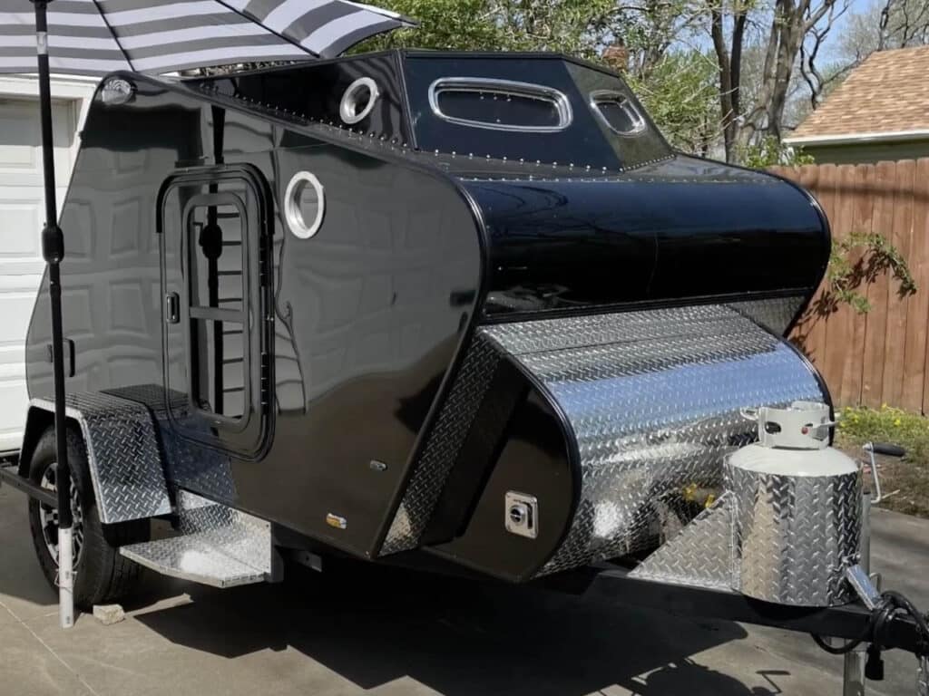 A sleek, compact camping trailer with a stylish design in black and aluminum, featuring circular windows that add an aesthetic touch. The trailer is equipped with sturdy tires and a robust entry platform.