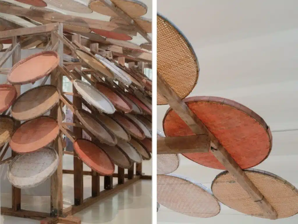 A collection of traditional handcrafted baskets made from natural materials, suspended on a multi-tiered wooden structure in an artistic, geometric arrangement. The baskets display natural colors and intricate designs, reflecting high craftsmanship and cultural ties to the local environment.