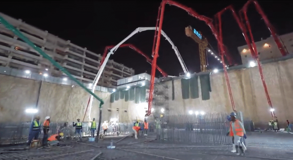Workers continue construction at night using a large concrete pump with an extended arm, under full site illumination.