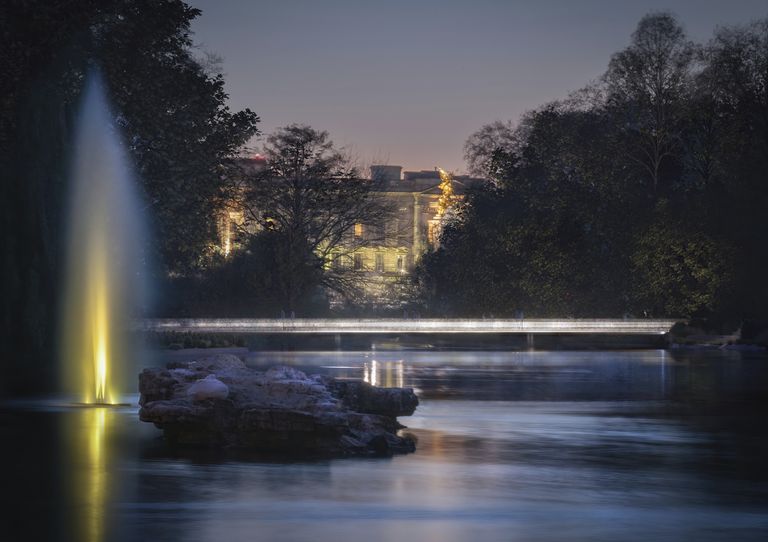 A lit bridge spanning over a river at night, with illuminated water jets shooting from the ground, surrounded by trees and backlit buildings glowing in the sky