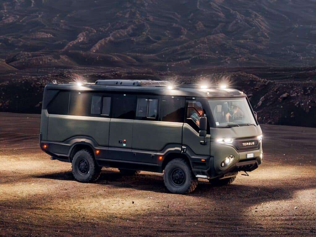 The Torsus Liberra bus drives on a desert road at night, with strong front lights illuminating the path against a stunning mountainous backdrop