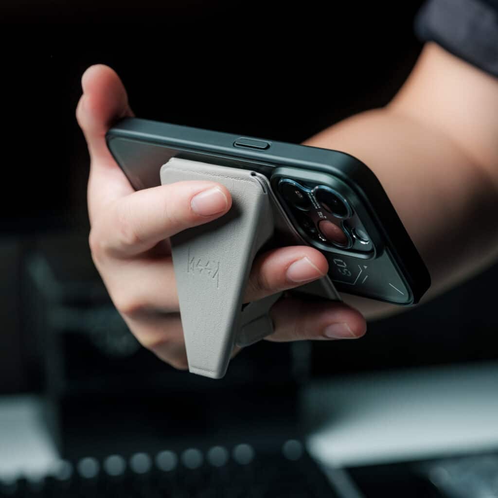 MagSafe Origami Grip Stand مثبت على iPhone، وهو في وضعية قائمة لعرض الفيديو، مع تصميم أنيق وأسلاك مغناطيسية.