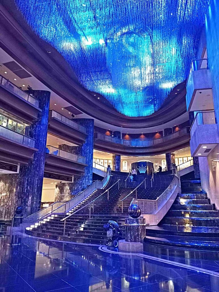 صورة داخلية لفضاء فاخر بتصميم مستقبلي، تظهر سلمًا متعرجًا مضاءً بإضاءة زرقاء، وسقف مزين بمصابيح متدلية تشبه الشلالات الضوئية، مع زوار يتجولون في المكان.
