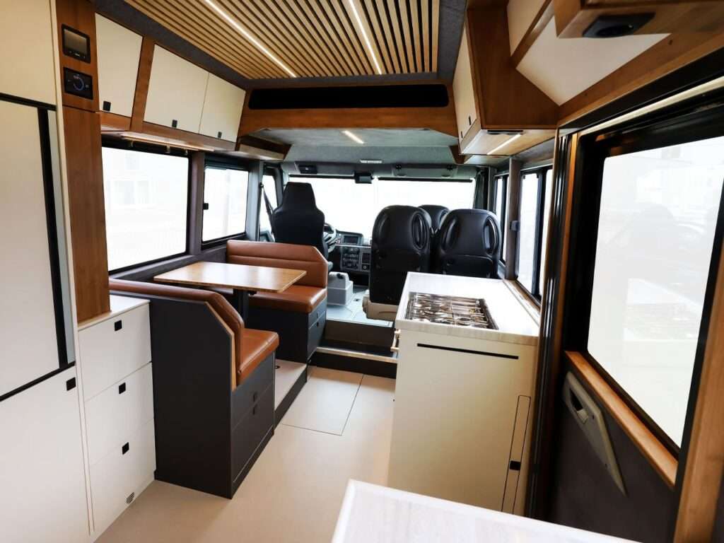 A modern kitchen in the Torsus Liberra bus, featuring integrated appliances like a stove and washing machine, with warm wooden lighting and built-in storage shelves, showcasing a functional yet luxurious design