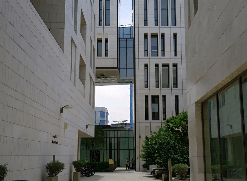 ممر ضيق بين مباني بيضاء ذات تصميم حديث، يُظهر نوافذ زجاجية كبيرة تسمح بدخول الضوء الطبيعي، مع وجود أشجار صغيرة وممرات مرصوفة تضيف لمسة من الطبيعة