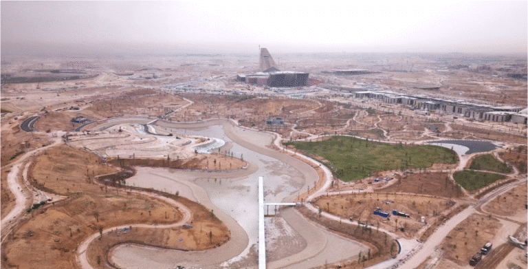 Aerial view showcasing the progress at King Salman Park in Riyadh, featuring winding paths surrounding growing green spaces and shimmering water features, with modern buildings emerging around the perimeter.