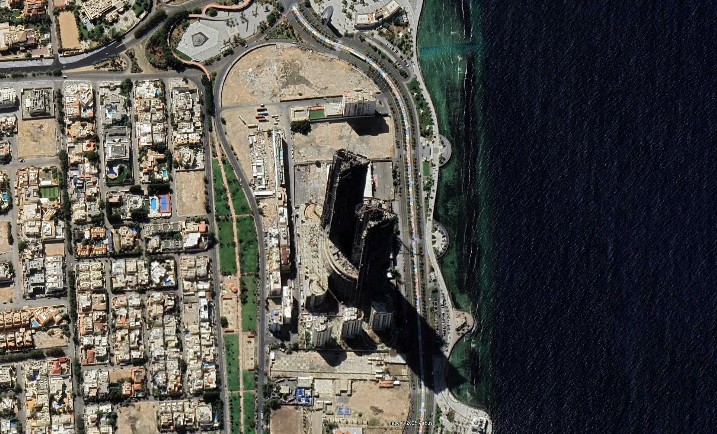 An aerial view of the Sama Towers in Jeddah, Saudi Arabia, showing the massive structures under construction near the waterfront. The area is surrounded by pedestrian pathways and trees, with residential buildings and roads visible in the background.