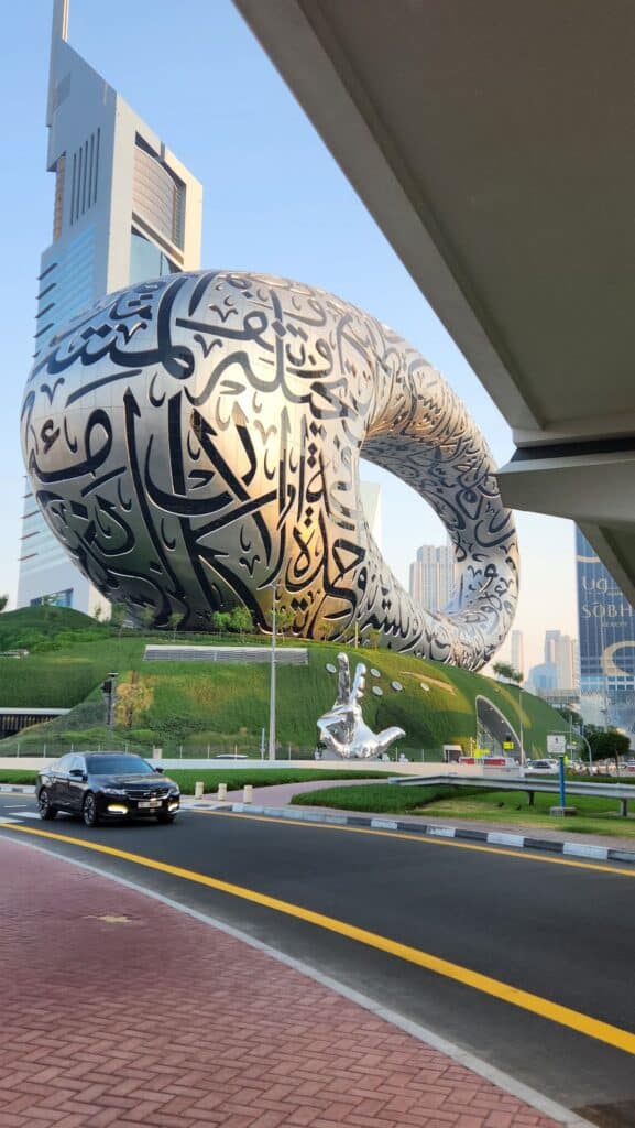 صورة نهارية لمتحف المستقبل في دبي من مستوى الشارع، يظهر تصميمه الحلقي المزين بخط عربي على تل أخضر، مع سيارة تسير على الطريق وناطحات سحاب في الخلفية.