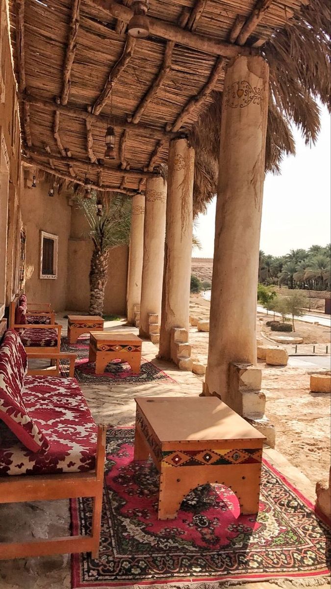 A traditional resting area in the desert, featuring wooden furniture that reflects local heritage, thick mud-brick columns, and a thatched roof, surrounded by palm trees and colorful rugs