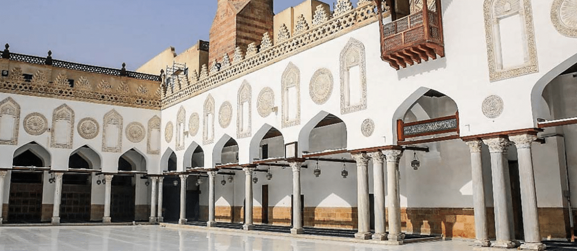 A traditional mosque facade featuring white arches, marble columns, and intricate botanical decorations with Arabic calligraphy on the walls.