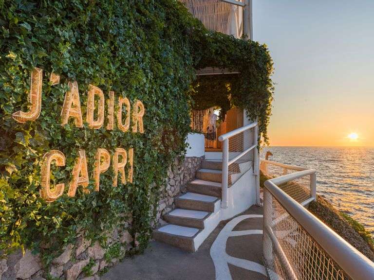 مدخل فندق يُزينه نباتات خضراء كثيفة، ويظهر عليه شعار 'J'Adior Capri' بإضاءة لافتة، مع إطلالة مباشرة على البحر في غروب الشمس.