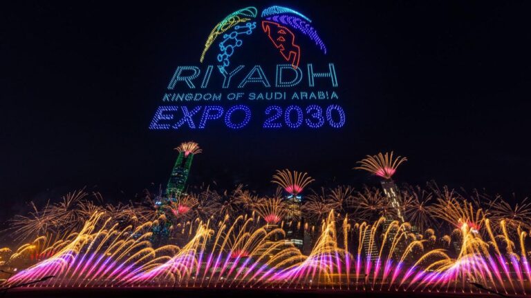 شعار معرض إكسبو 2030 الرياض، مضاء بأضواء ليد ملونة، مع عروض ألعاب نارية خلفه تضيء السماء.