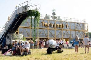 A group of visitors sitting and standing in front of the 'Terminal 1' space at Glastonbury Festival, with a large sign reading 'Terminal 1' visible, surrounded by festive energy and movement