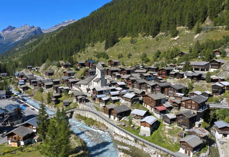 A small Swiss village nestled between mountains, built on green slopes with a small river flowing through it, featuring traditional wooden houses and a central church.