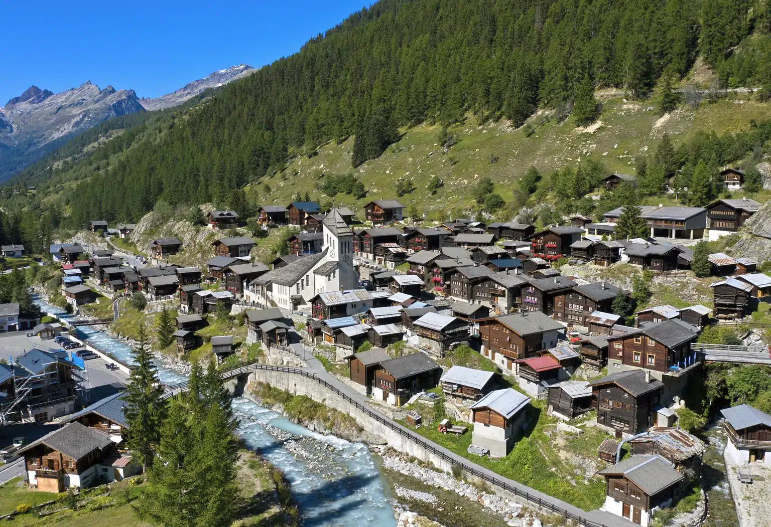 A small Swiss village nestled between mountains, built on green slopes with a small river flowing through it, featuring traditional wooden houses and a central church.