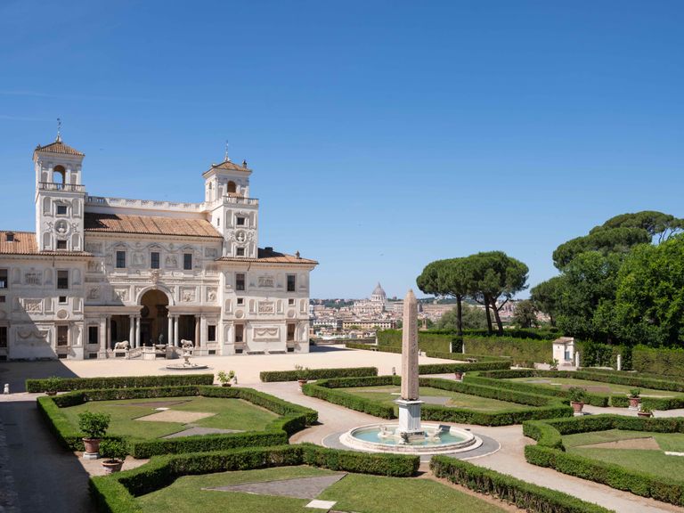 Villa Medici in Rome, showcasing the elegant villa surrounded by meticulously landscaped gardens, featuring a central fountain and a tall obelisk. The background reveals a stunning panoramic view of Rome, including the iconic Vatican City.