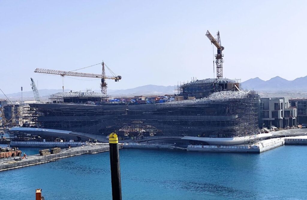 A large-scale construction site with multiple cranes and scaffolding surrounding a modern building under development. The structure is situated near the water, with clear blue skies and distant mountains in the background. The scene captures the early stages of architectural progress, showcasing the integration of design with the natural environment.