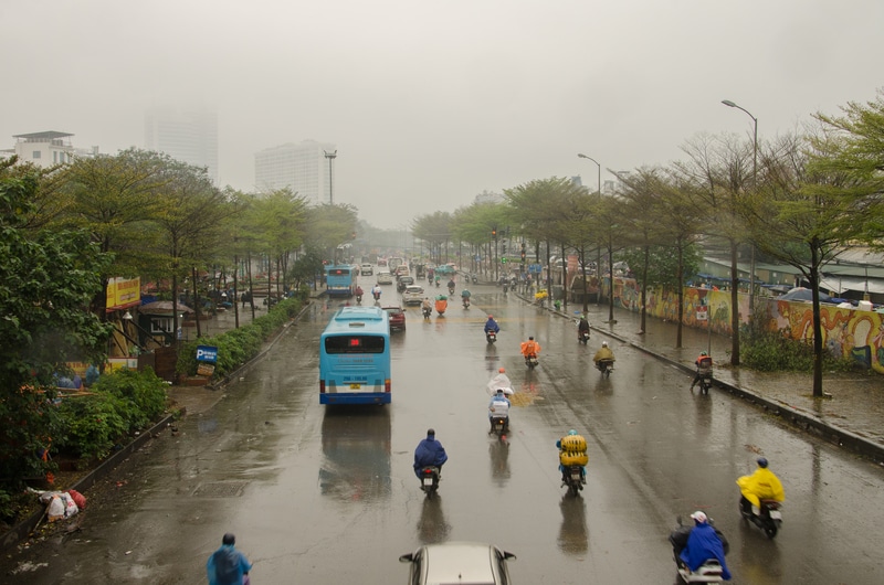 A rainy street in Hanoi with numerous motorbikes and cars, featuring a large bus on the road