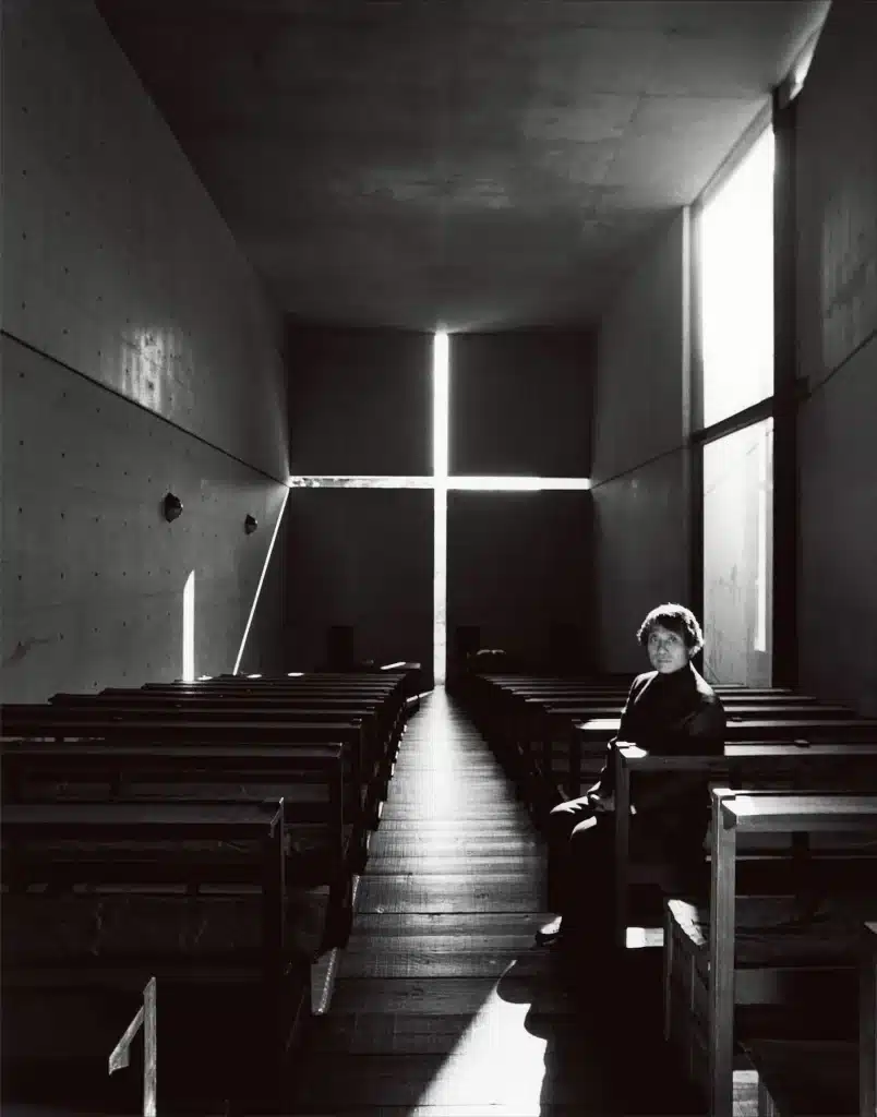 Natural light slicing through concrete in a cross shape, highlighting simple wooden pews.