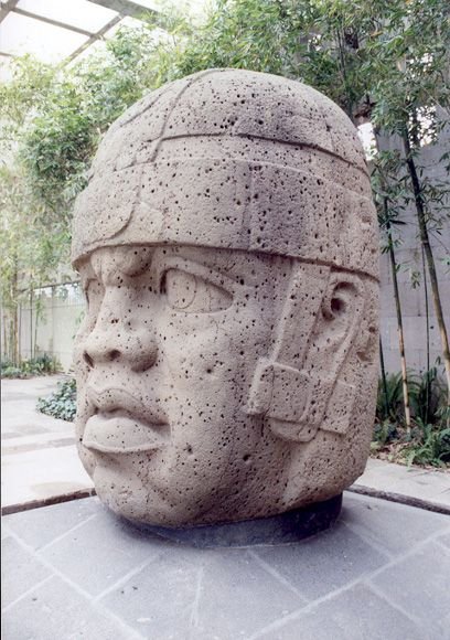 A side view of an Olmec head inside a museum, showing its massive scale compared to surrounding visitors.
