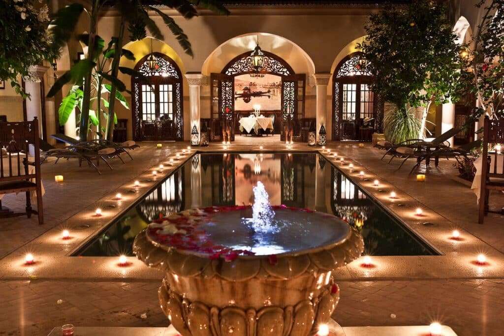 An indoor garden in a Moroccan riad featuring a fountain surrounded by lush plants and colorful mosaic tiles.