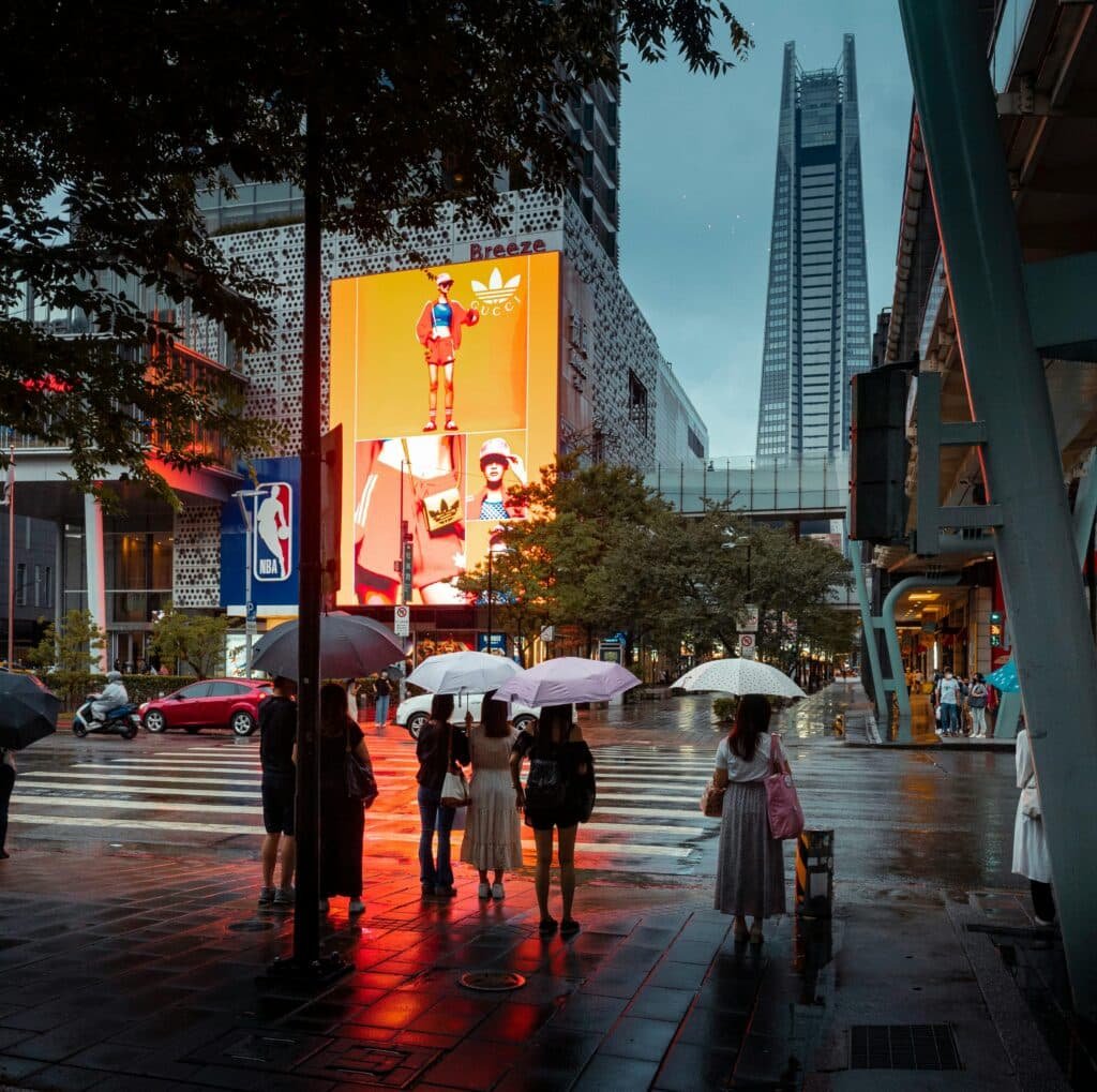 A group of people standing under umbrellas at a city intersection lit by large commercial billboards on a rainy day in a modern urban setting.
