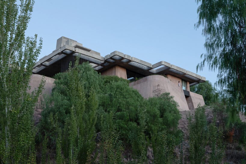 الجزء الجانبي للمنزل يظهر كيف يندمج الهيكل الخرساني مع الأشجار والنباتات الطبيعية المحيطة، مع وجود نوافذ عريضة تسمح بدخول الضوء.