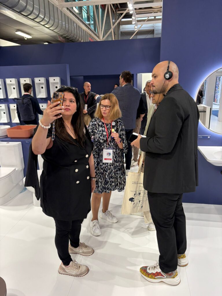 مجموعة من المتخصصين في التصميم يتبادلون الحديث في جناح بمعرض Cersaie 2025، محاطين بأحدث منتجات الحمامات. إحدى النساء تلتقط صورة بهاتفها بينما تحمل أخرى ميكروفونًا، مما يشير إلى جولة إرشادية أو مقابلة. المشهد يعكس تركيز المعرض على الابتكار المعماري والحرفية الإيطالية.