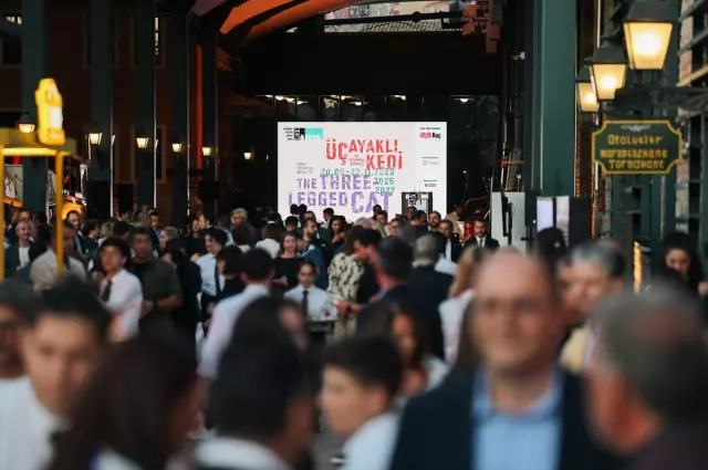 A bustling crowd gathers under the arched entrance of a historic building during the opening of the 18th Istanbul Biennial, with a large banner reading &ldquo;&Uuml;&ccedil;ayaklı Kedi / The Three-Legged Cat&rdquo; visible in the background.