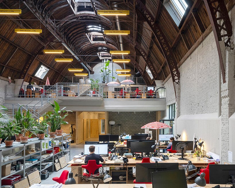 A spacious architectural studio housed in a renovated historic building, featuring a curved wooden ceiling, high skylights, and exposed structural elements. The upper level includes a mezzanine workspace connected by metal stairs, while the ground floor is filled with desks, computers, and indoor plants that bring life and warmth to the creative environment.