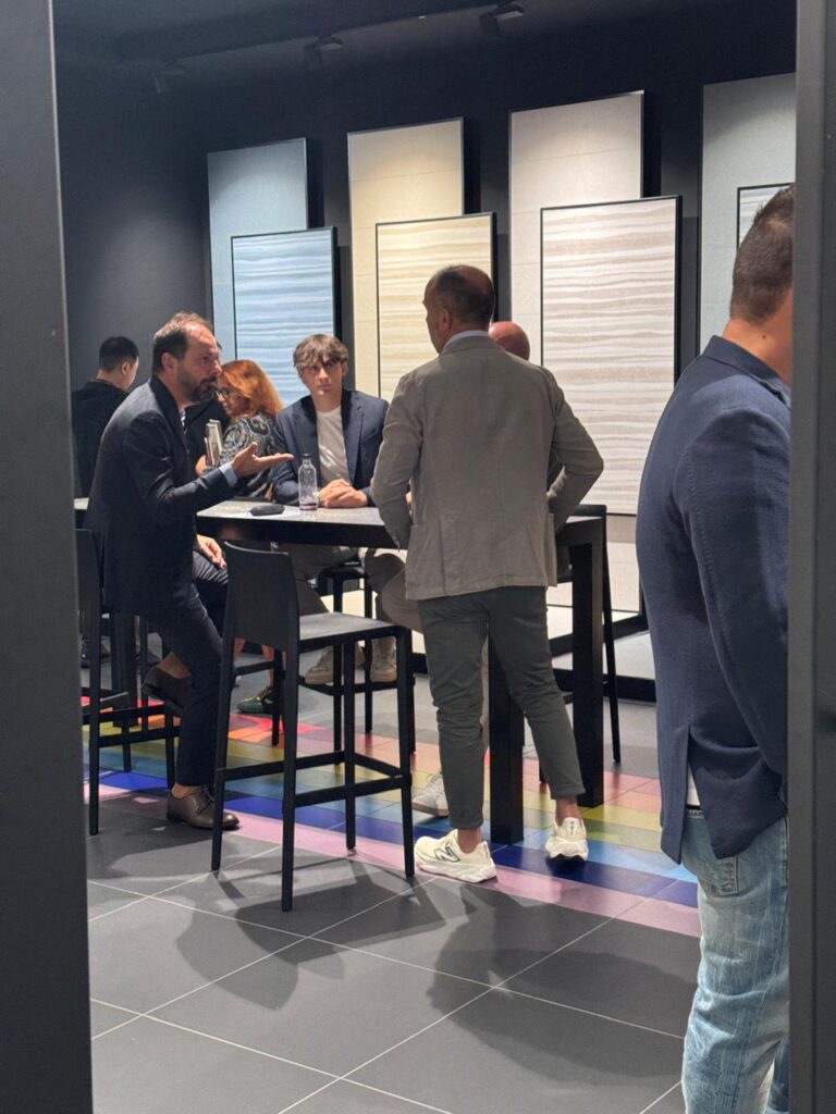 مجموعة من المشاركين في معرض Cersaie 2025 يتجاذبون أطراف الحديث حول طاولة في جناح عرض، مع خلفية تُظهر لوحات سيراميك بألوان هادئة وتصميمات عصرية، وسط أرضية رمادية تعكس الإضاءة الدافئة.