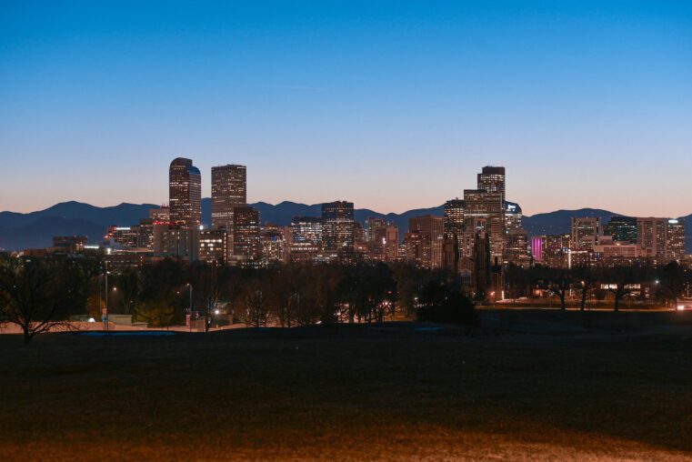 منظر ليلًا لمدينة دنفر مع أضواء المباني الشاهقة وخلفية الجبال، تُظهر جمال العمارة الحديثة والطبيعة المحيطة.