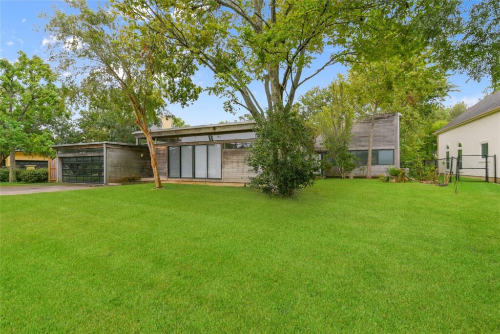 Victor Lundy&rsquo;s modernist home in Bellaire, featuring wood-paneled walls, large glass windows, and a lush green lawn surrounded by mature trees under a bright blue sky.