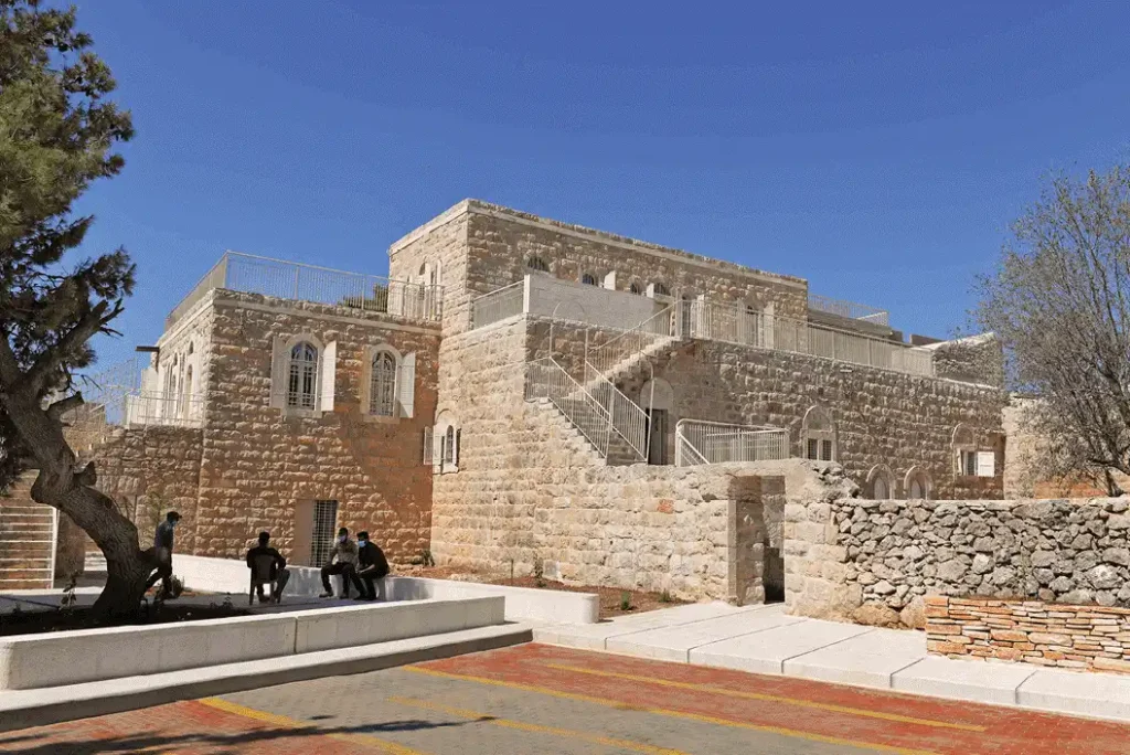 A restored traditional stone building with arched windows and modern white railings, set against a clear blue sky. People sit on a low concrete bench in the foreground, surrounded by trees and paved walkways.