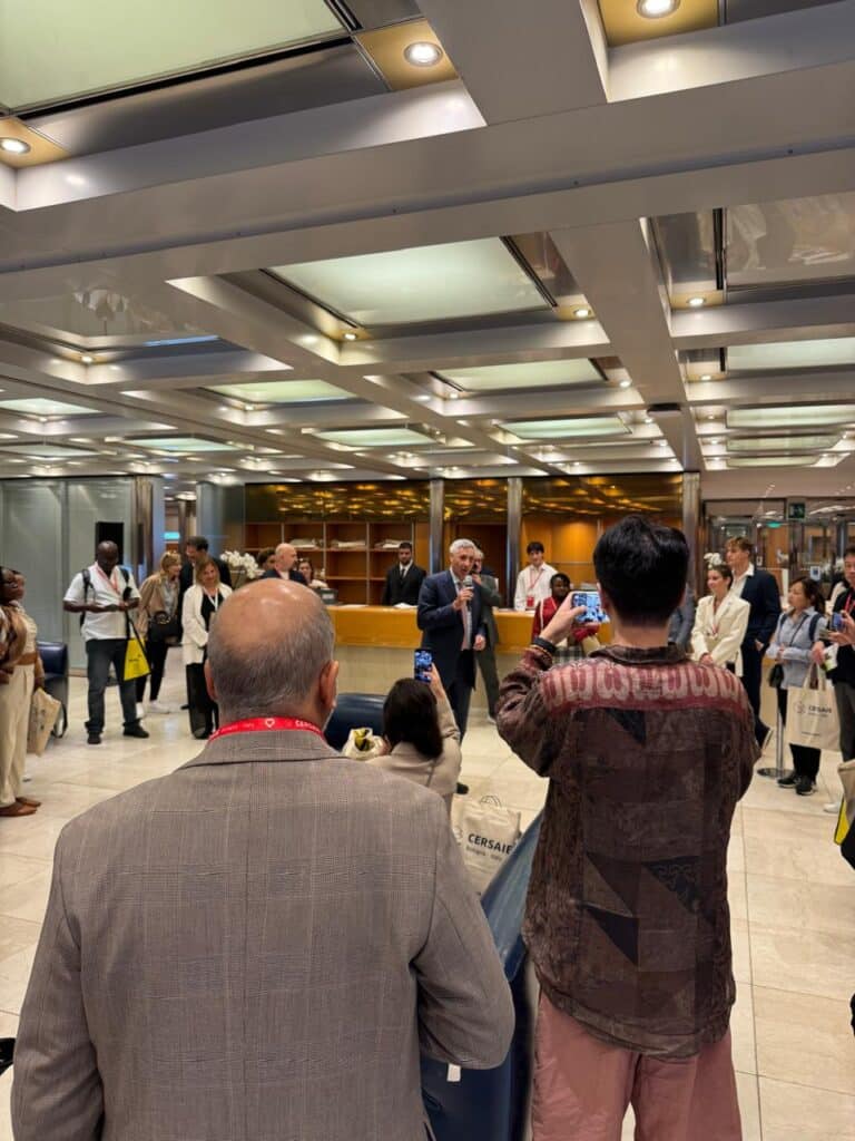 A speaker addresses a small, attentive audience at Cersaie 2025&rsquo;s welcome area. Attendees, some recording on phones, listen as he speaks near the reception desk &mdash; a moment of connection before diving into the fair&rsquo;s design wonders.